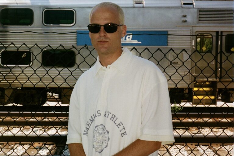 Devault in dark sunglasses and a cream polo shirt standing behind a chain-link fence with a silver commuter train on the tracks behind him