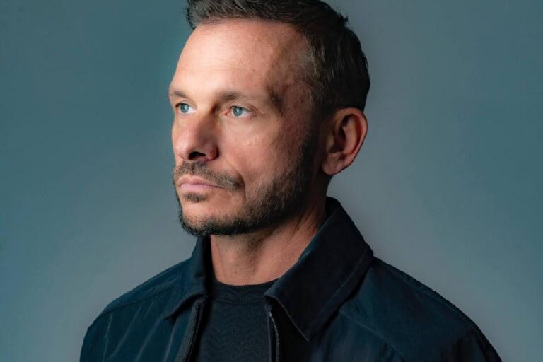 Andy C press portrait, looking off-camera in a dark jacket against a grey backdrop