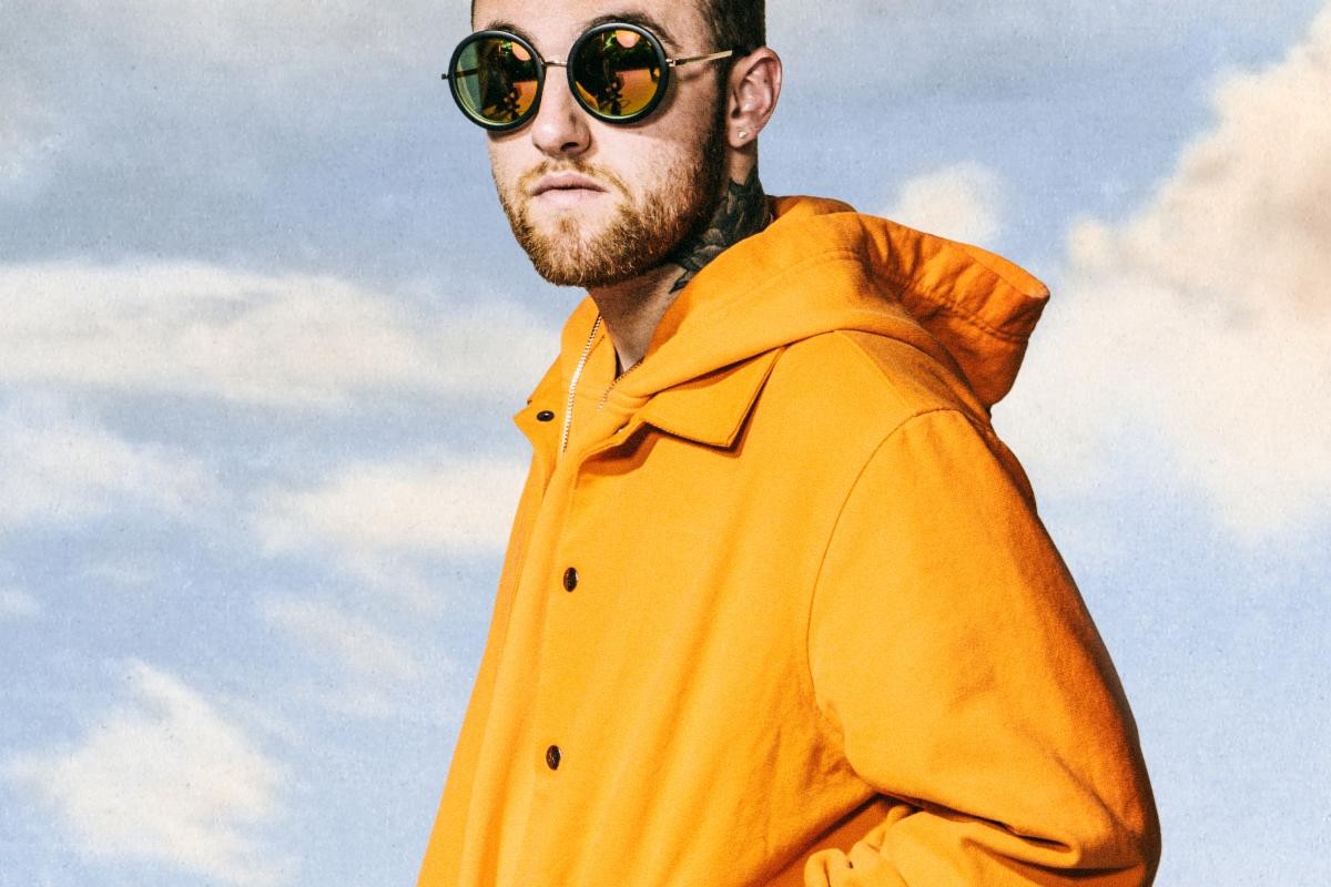 Mac Miller in a bright orange hooded jacket and round mirrored sunglasses, photographed against a pale blue cloudy sky