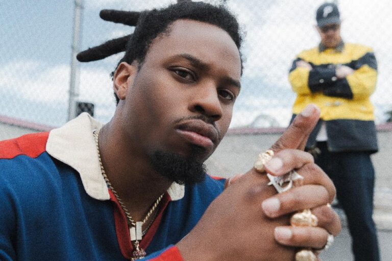 Denzel Curry leaning toward the camera in a blue and red rugby shirt with gold chains and rings, a figure in a yellow jacket standing behind him beside a chain-link fence