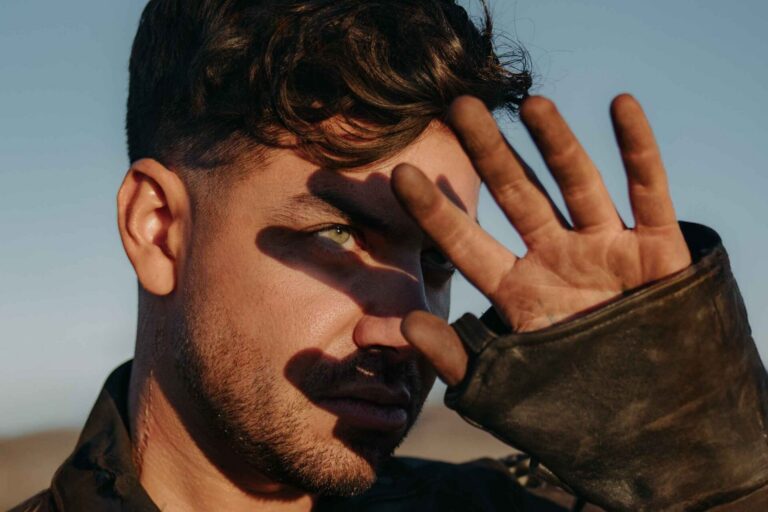 Close-up press portrait of Anyma raising a hand to shade his eyes from low sun, wearing a weathered leather jacket against a pale blue sky