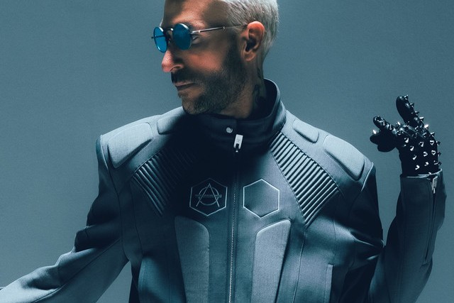 Don Diablo press portrait in a grey armored jacket with the HEXAGON logo, blue round sunglasses and a studded black glove against a grey studio backdrop