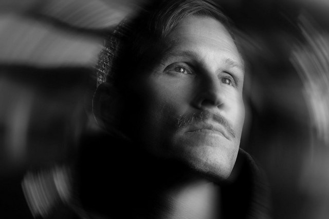 Black and white press portrait of Kaskade looking upward, his face lit against a dark background framed by streaks of motion blur