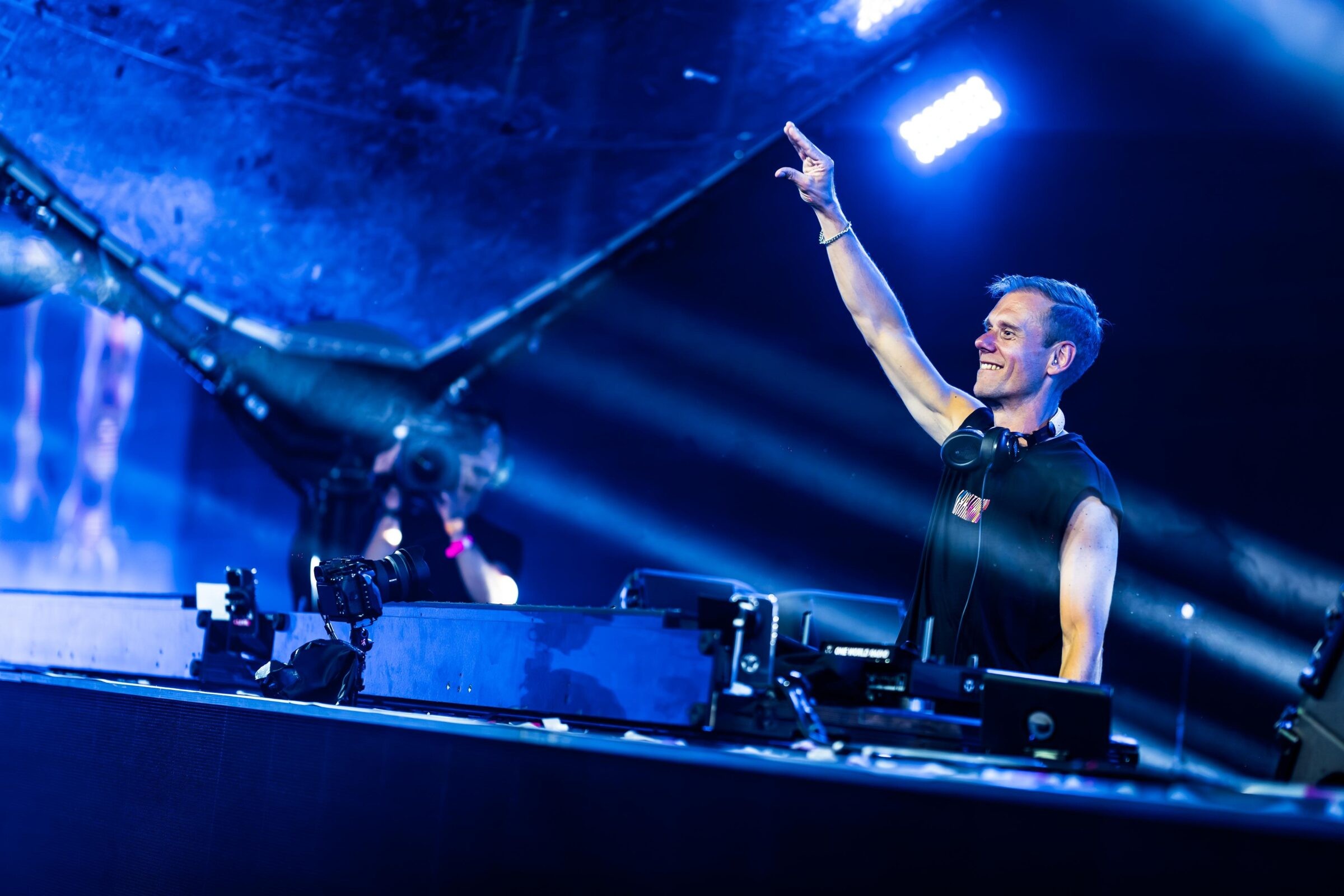 Armin van Buuren smiles and points to the crowd from behind the DJ decks at a Tomorrowland Mainstage set, lit in blue stage light