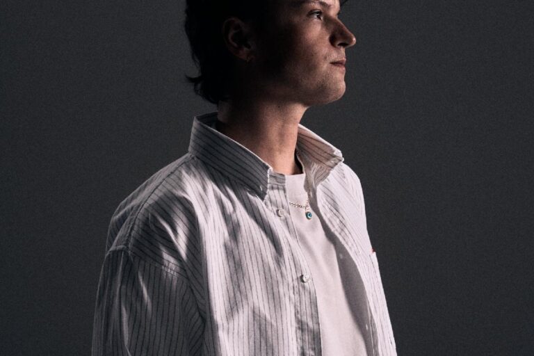 MPH press portrait in an oversized white pinstriped shirt, looking off camera against a dark grey backdrop