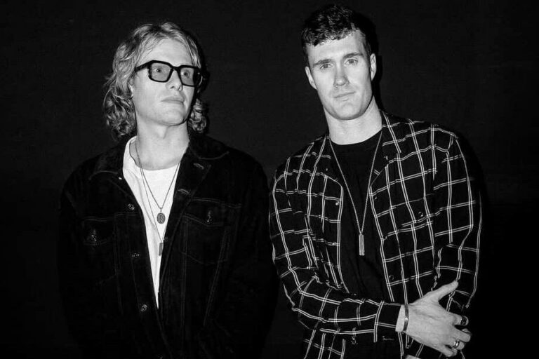 Bob Moses duo Tom Howie and Jimmy Vallance in a black-and-white press portrait, one in dark-framed glasses and a denim jacket, the other in a windowpane-check shirt, against a black backdrop