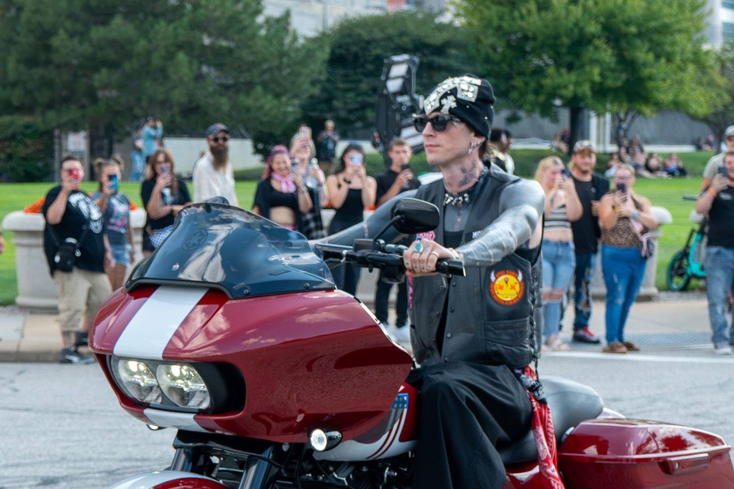 mgk in sunglasses, a jewelled black beanie and a leather vest riding a red and white Harley-Davidson through Cleveland on the MGK Day City Ride, with fans filming from the sidewalk