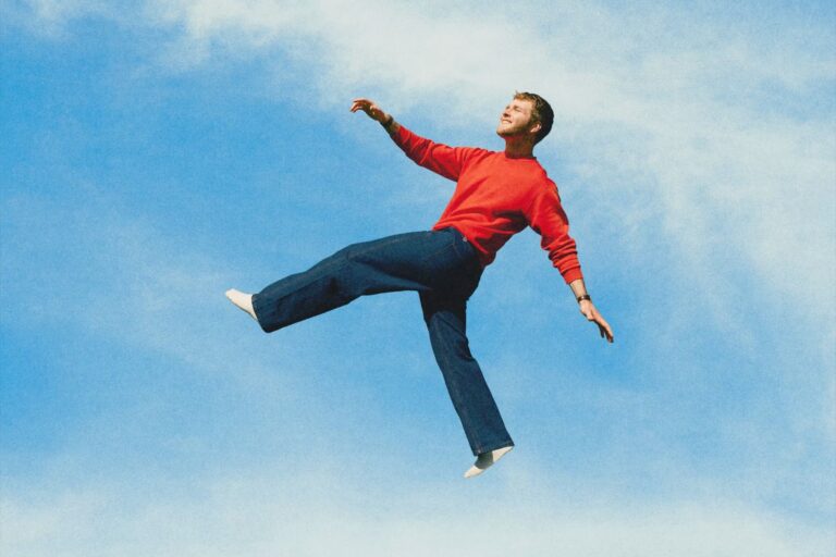 Alex Warren suspended mid-air against a bright blue sky in a red sweatshirt and dark blue jeans, from the WILDCHILD album cover