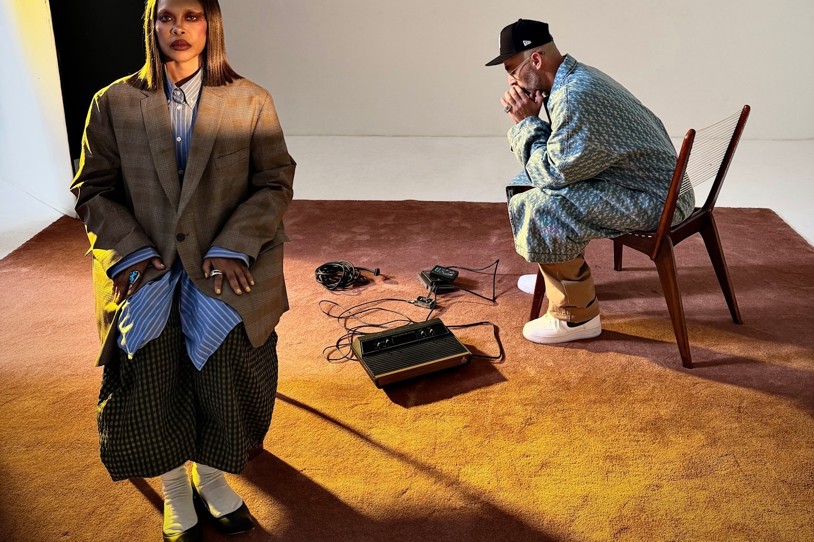 Erykah Badu stands in an oversized checked blazer while The Alchemist sits on a wooden chair, a vintage Atari console and tangled cables on the rust carpet between them