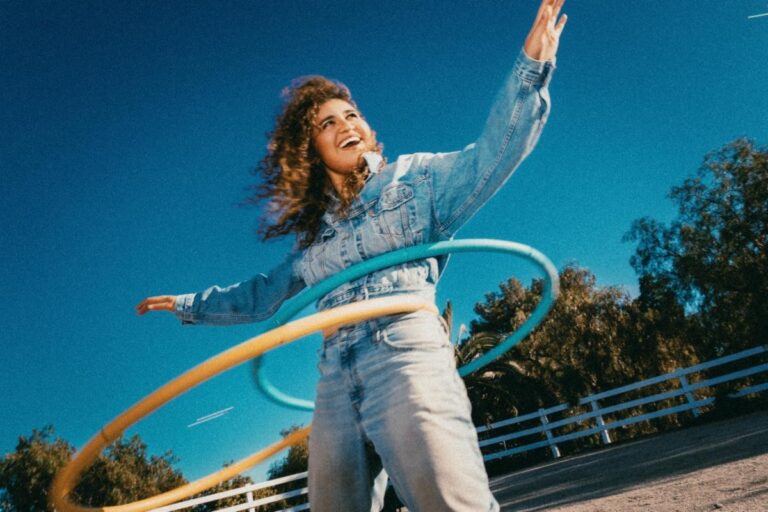 Remi Wolf laughing in a denim jacket and jeans, spinning two hula hoops around her waist against a deep blue sky
