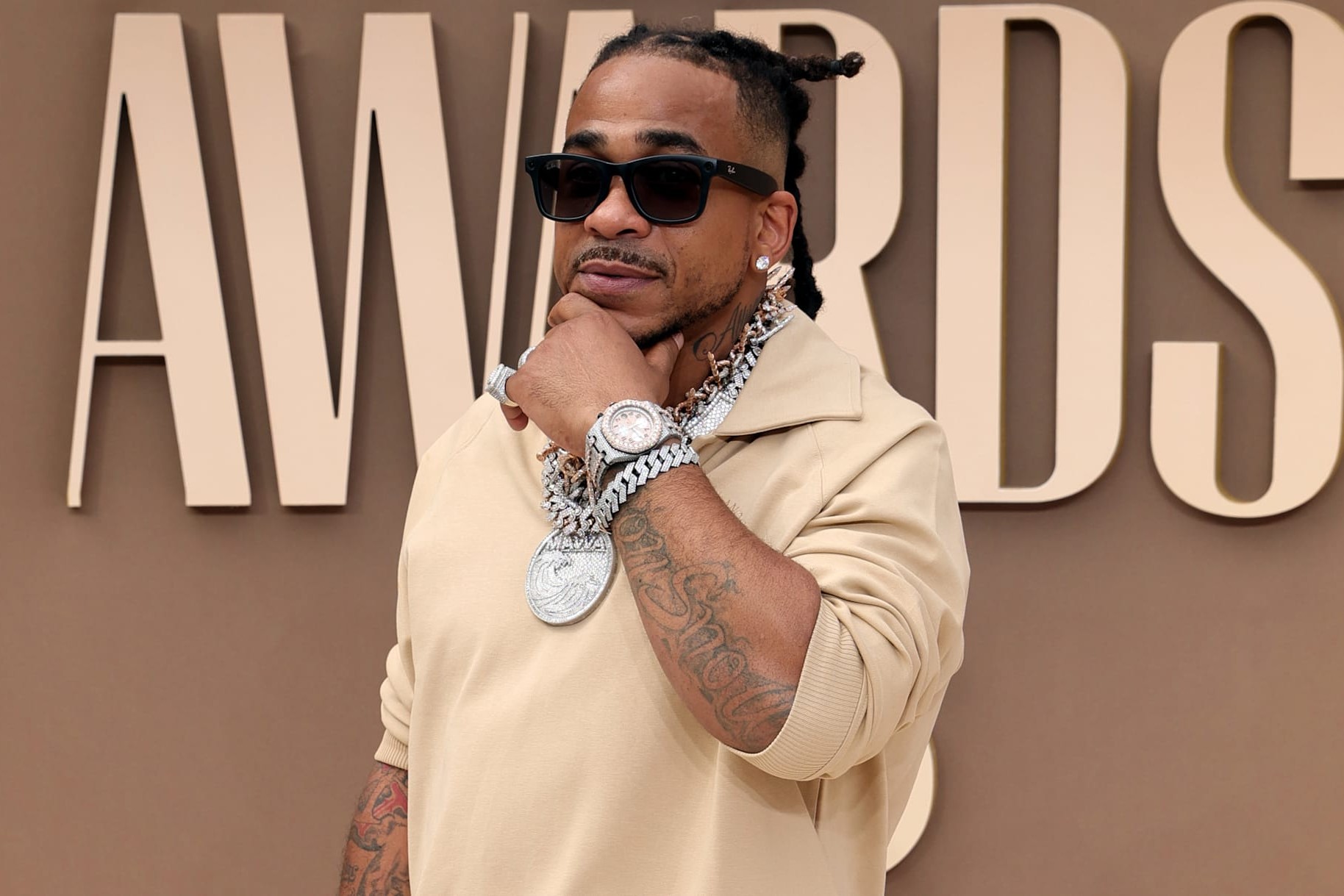 Max B poses on the BET Awards 2026 press-room carpet in a tan trench coat, sunglasses, and heavy diamond chains, hand resting under his chin