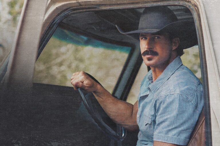 Riley Green in a black cowboy hat and blue striped western shirt sitting behind the wheel of a vintage pickup truck, looking out through the open window