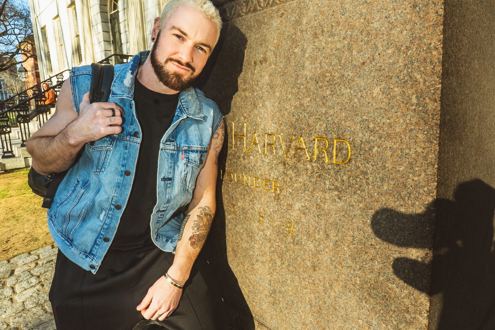 James Hype, in a denim vest and black outfit, leans against the Harvard founder statue base on the Harvard campus