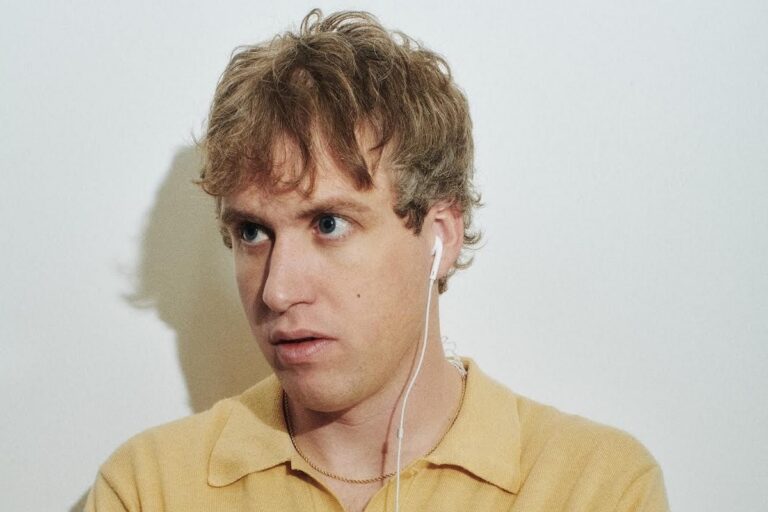 jigitz in a pale yellow polo shirt with white wired earbuds in, looking off to the side against a plain white wall