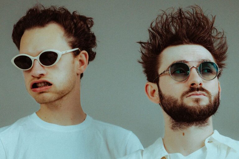 The two members of Louis The Child side by side in white shirts and sunglasses against a grey studio backdrop