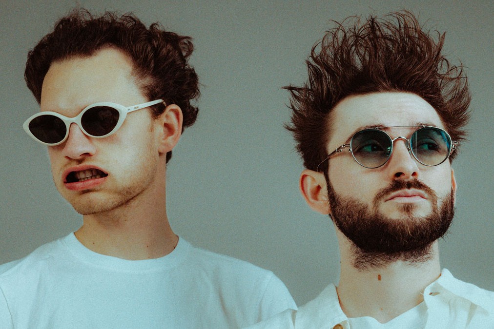 The two members of Louis The Child side by side in white shirts and sunglasses against a grey studio backdrop