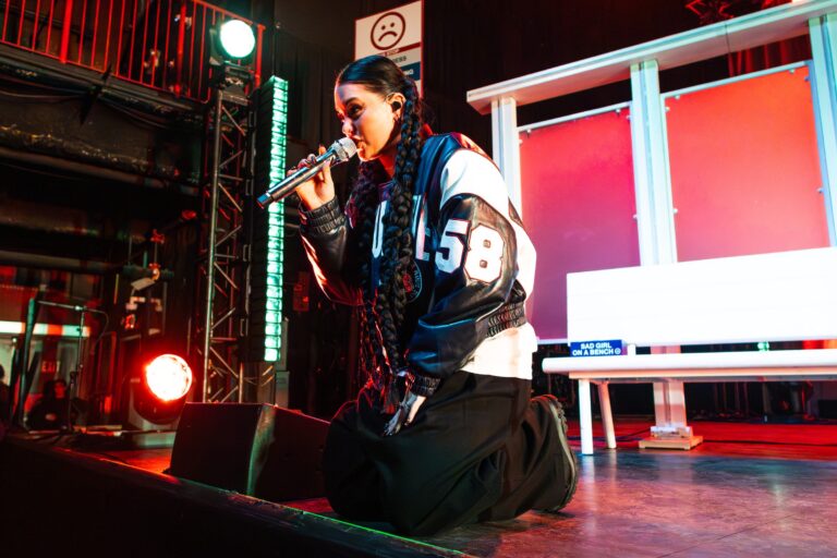 Lauren Spencer Smith kneels onstage singing into a microphone in a black-and-white varsity jacket during her Art of Being a Mess Tour stop in Boston