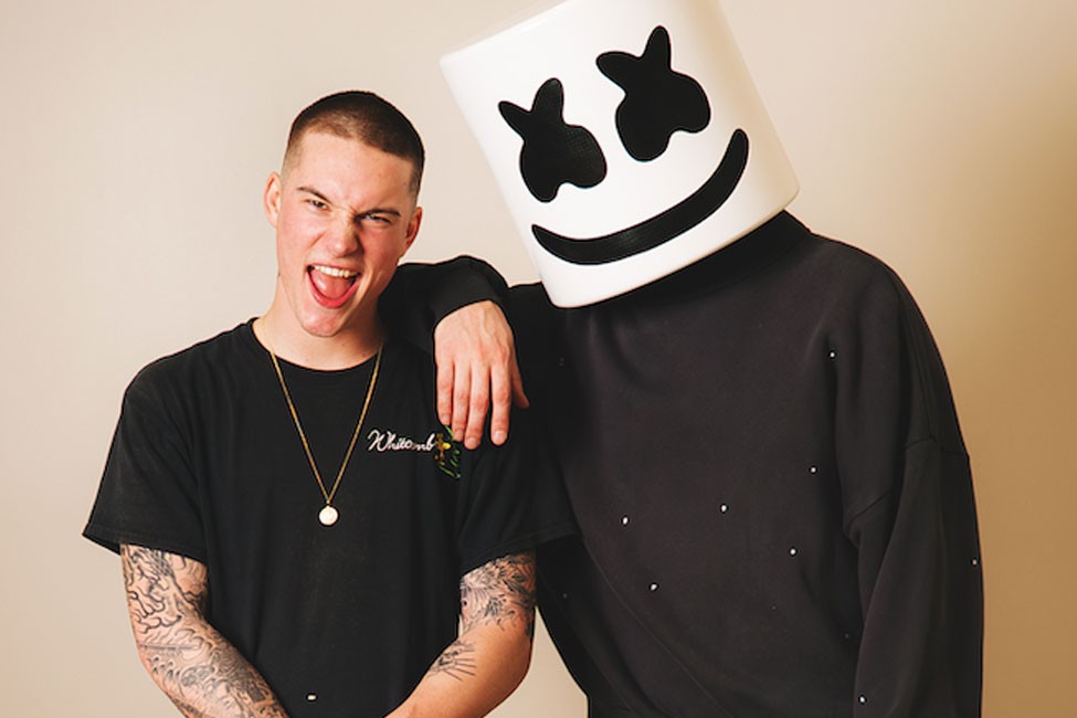 Marshmello in his white helmet and black sweatshirt resting an arm on Cameron Whitcomb, who wears a black tee and gold chain and pulls a face, against a cream backdrop
