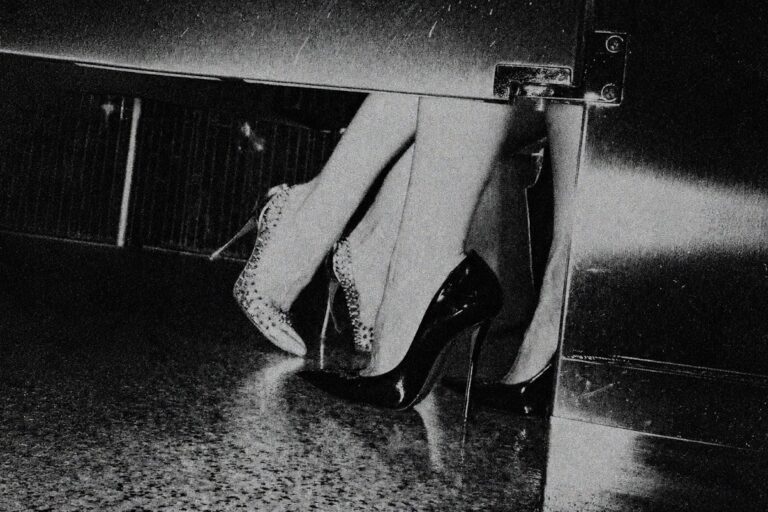 Grainy black and white key art from the Long Jacket video showing a pair of legs in studded and patent stiletto heels beside a metal door on a wet reflective floor