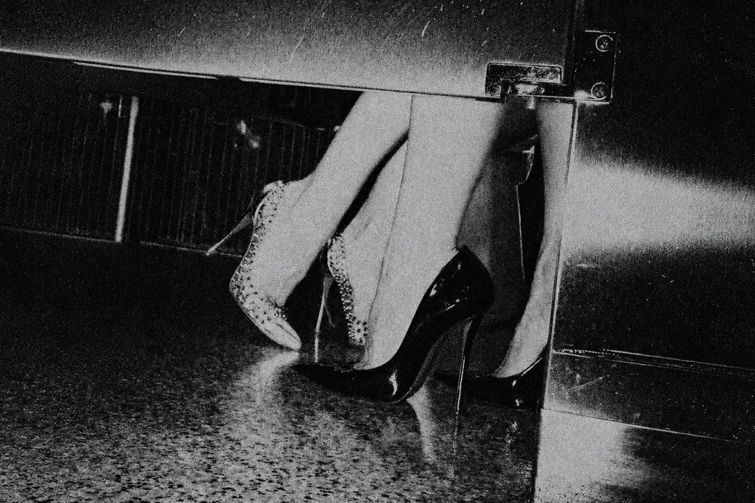 Grainy black and white key art from the Long Jacket video showing a pair of legs in studded and patent stiletto heels beside a metal door on a wet reflective floor
