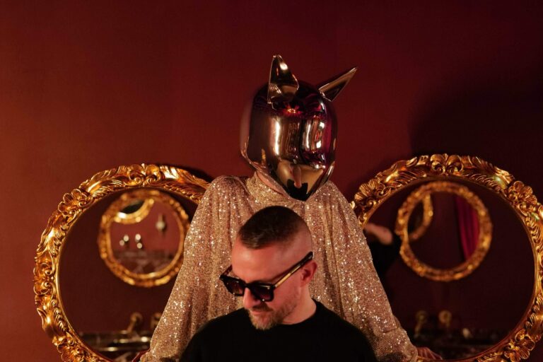 Galantis in sunglasses and a black tee in front of a figure wearing a chrome cat-eared mask and gold sequined robe, flanked by two ornate gold mirror frames against a deep red wall