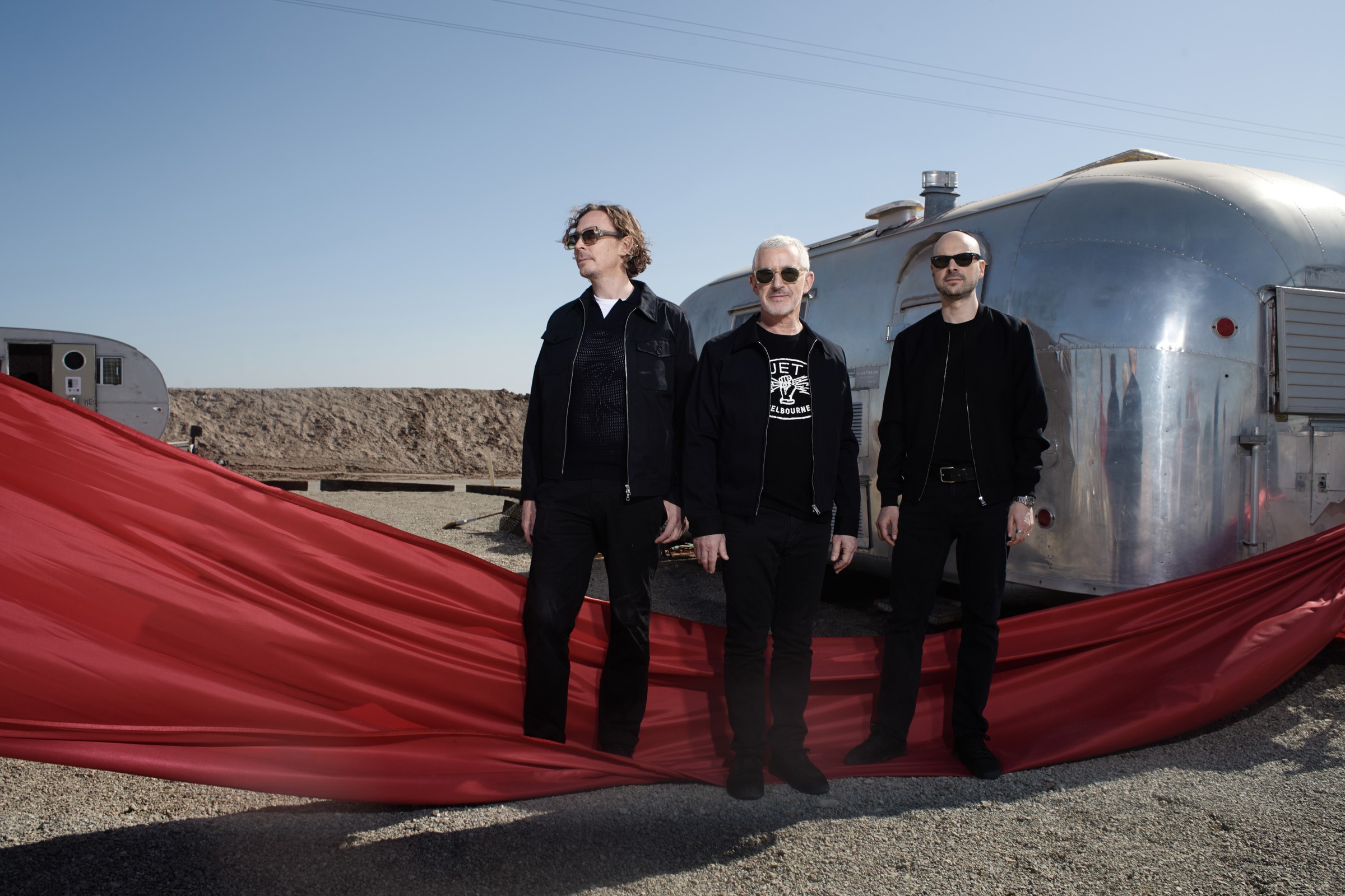 Above & Beyond press photo: the three members in black standing on desert gravel beside a silver Airstream trailer, with a long red fabric train sweeping across the ground in front of them