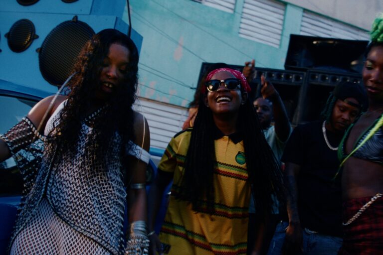 Still from the Send Shots video showing Original Koffee in a yellow Jamaica-colours polo and red bandana laughing among dancers in front of a sound system speaker stack on a Kingston street