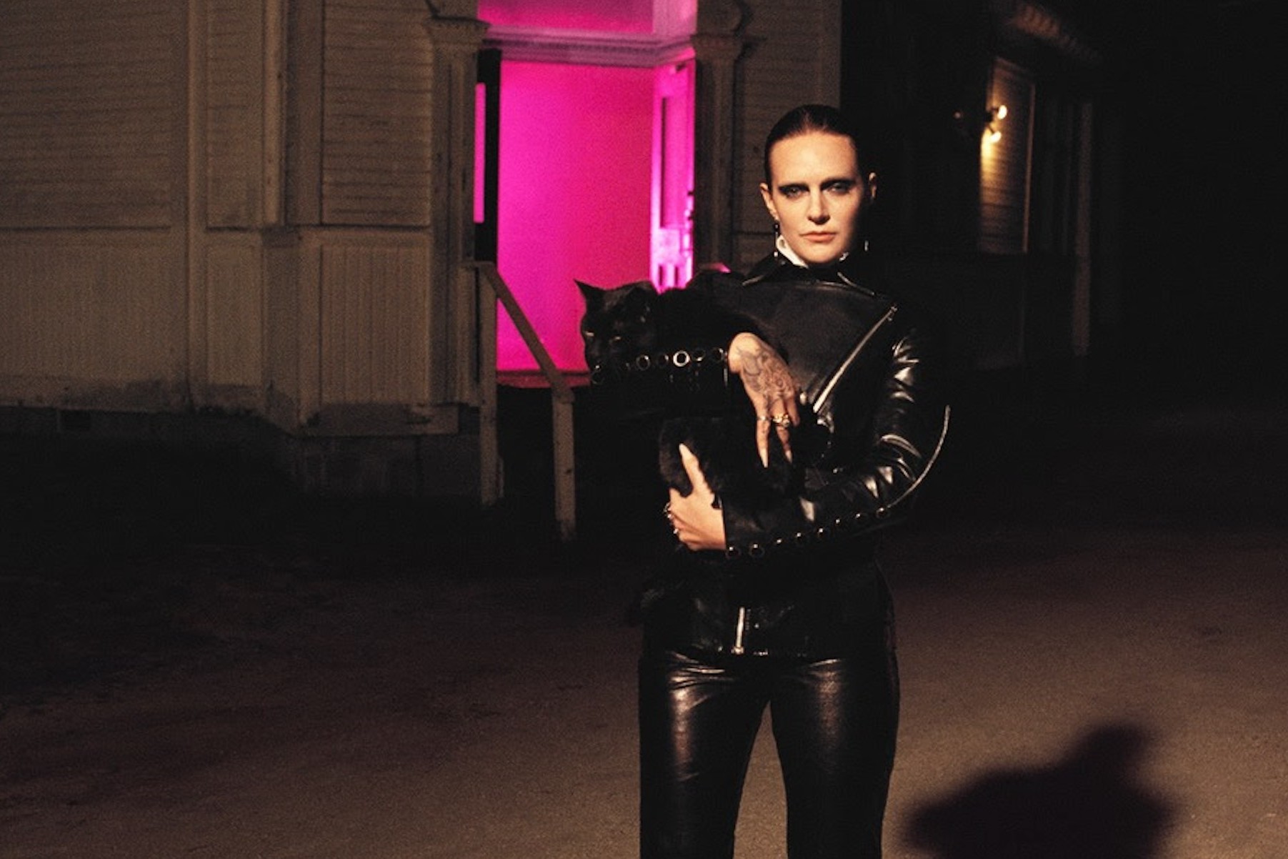 Tove Lo stands in a doorway lit by pink light at night, dressed in black leather and holding a black cat, in a press photo for her single DNH