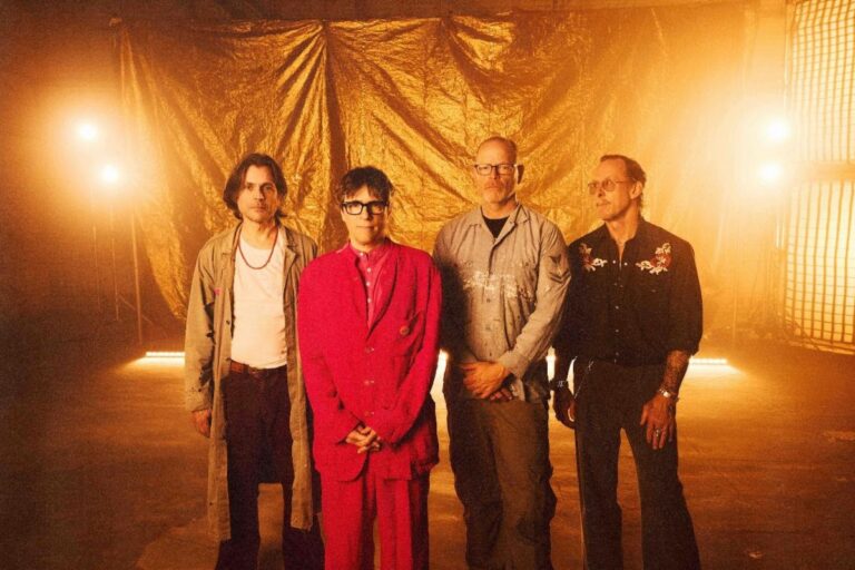 The four members of Weezer standing in a row in front of a crumpled gold backdrop under warm stage lights, Rivers Cuomo center in a red suit