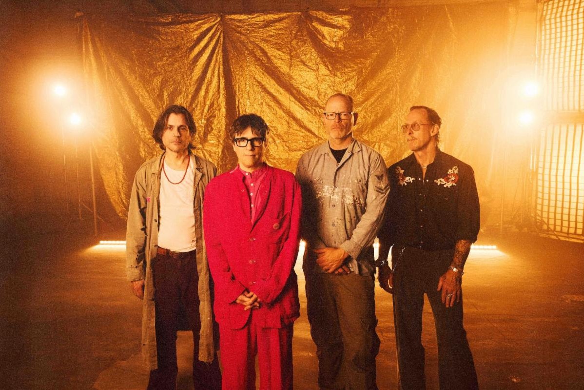 The four members of Weezer standing in a row in front of a crumpled gold backdrop under warm stage lights, Rivers Cuomo center in a red suit