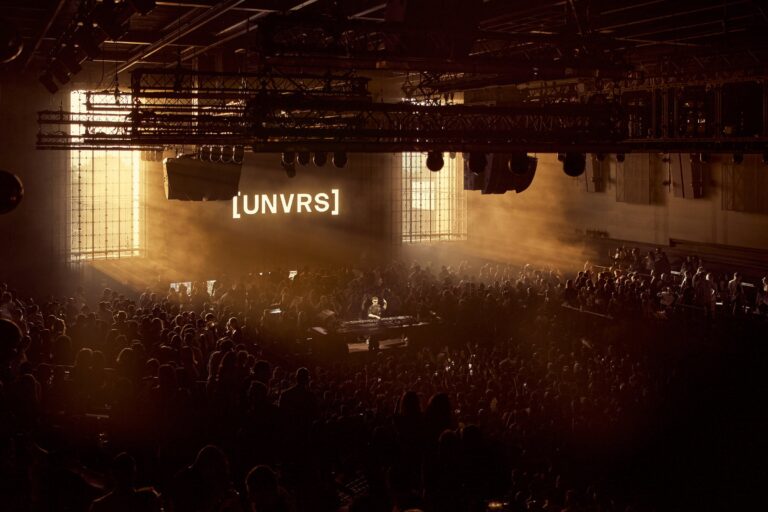 Packed dark dancefloor at [UNVRS] Ibiza lit by golden haze, with a DJ at the booth below a large illuminated [UNVRS] wordmark and overhead lighting truss