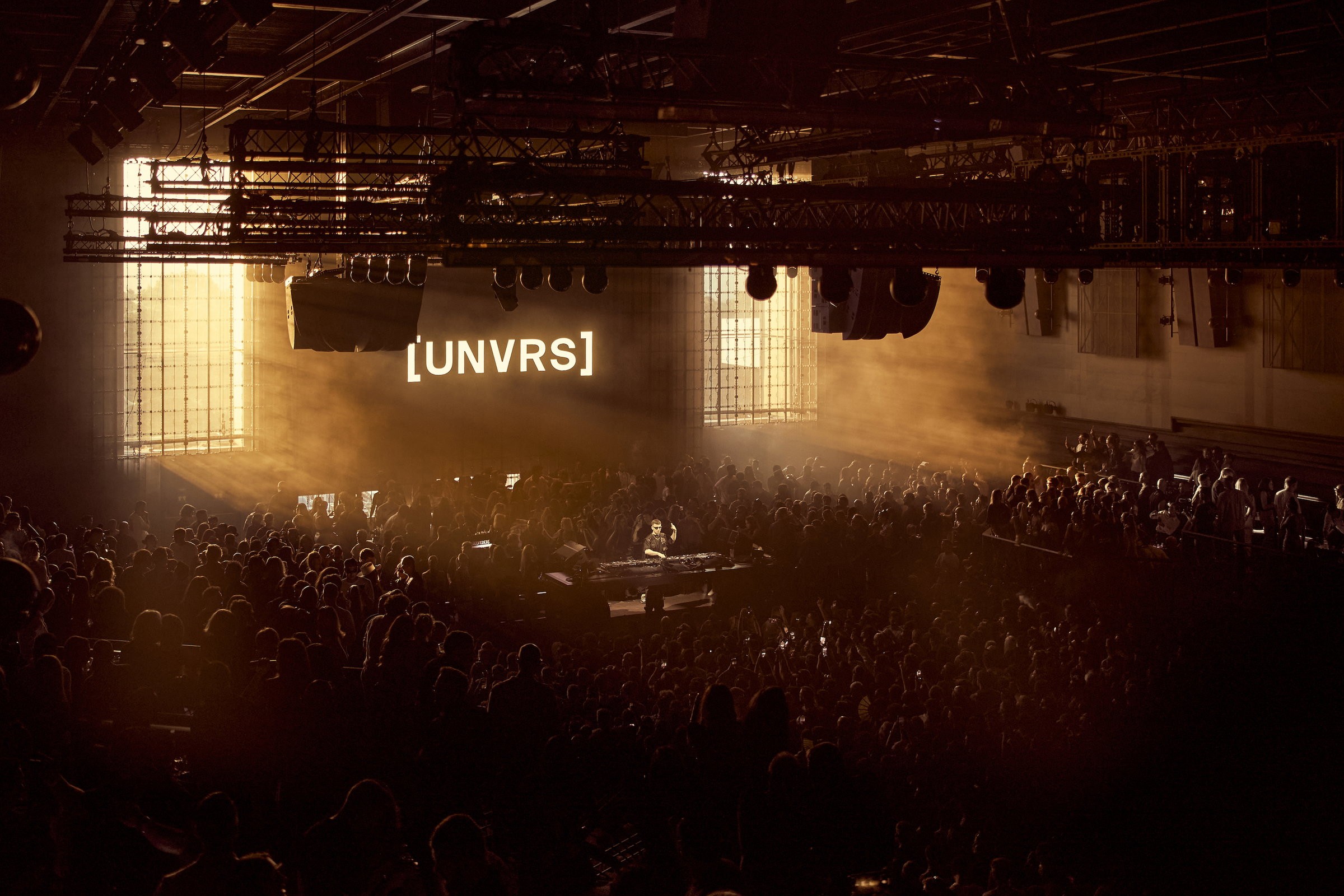 Packed dark dancefloor at [UNVRS] Ibiza lit by golden haze, with a DJ at the booth below a large illuminated [UNVRS] wordmark and overhead lighting truss