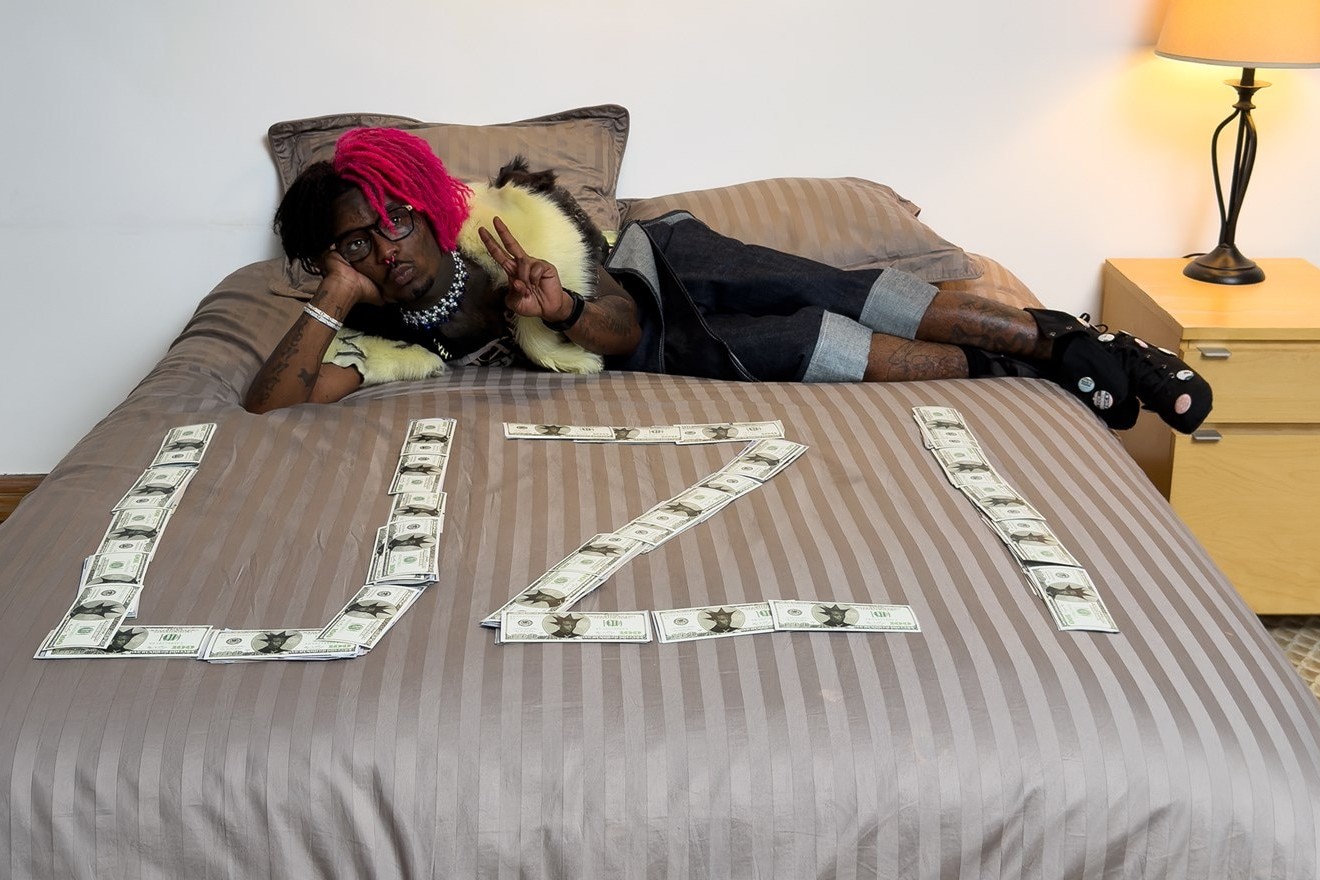 Lil Uzi Vert, with pink dreadlocks and glasses, lies on a bed flashing a peace sign next to cash spelling out UZI, from a Dazed magazine cover shoot