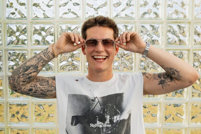 Cameron Whitcomb smiles while putting on sunglasses in front of a glass-block wall, wearing a Rolling Stone t-shirt, from his Rolling Stone Canada cover story