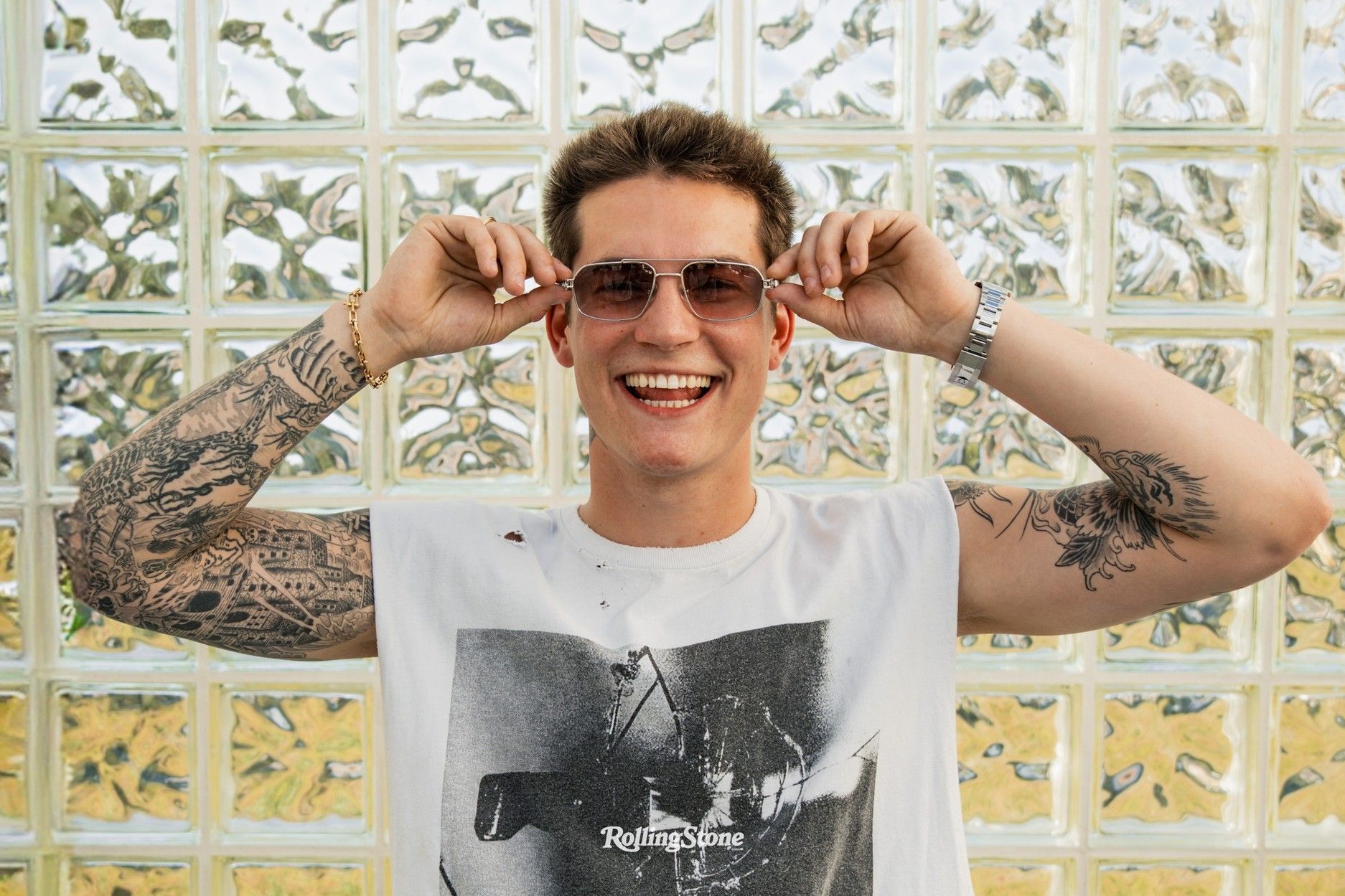 Cameron Whitcomb smiles while putting on sunglasses in front of a glass-block wall, wearing a Rolling Stone t-shirt, from his Rolling Stone Canada cover story