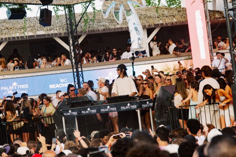 A DJ plays to a packed daytime crowd at Beachclub, with a PALM TREE Montreal LED wall, a BEACHCLUB logo and a mirrored palm-tree sculpture under a thatched roof