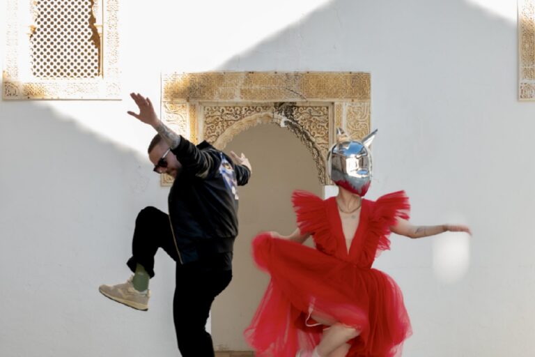 Galantis and Piiper leaping in front of a whitewashed wall with a carved Moorish arch, him in a black leather bomber and sunglasses mid-kick, her spinning in a red ruffled tulle dress and a mirrored chrome animal mask