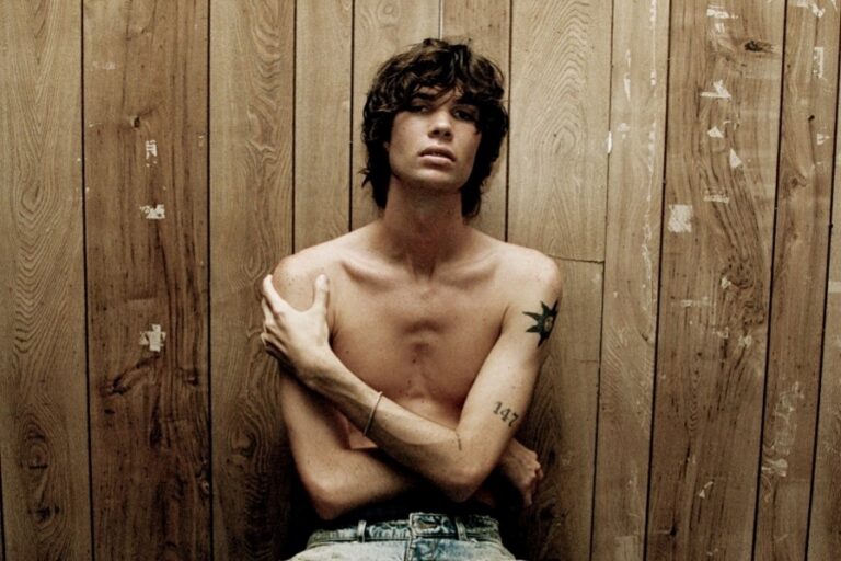 Will Swinton press portrait, shirtless with a star tattoo on his chest and arms folded, seated against a weathered wood plank wall