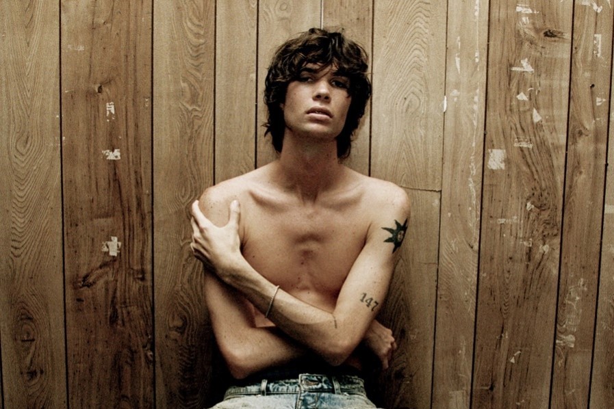 Will Swinton press portrait, shirtless with a star tattoo on his chest and arms folded, seated against a weathered wood plank wall