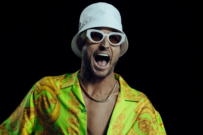 FISHER in a white bucket hat, white crystal-studded sunglasses and an open green-and-gold baroque-print silk shirt with a gold chain, mouth open mid-shout against a black backdrop