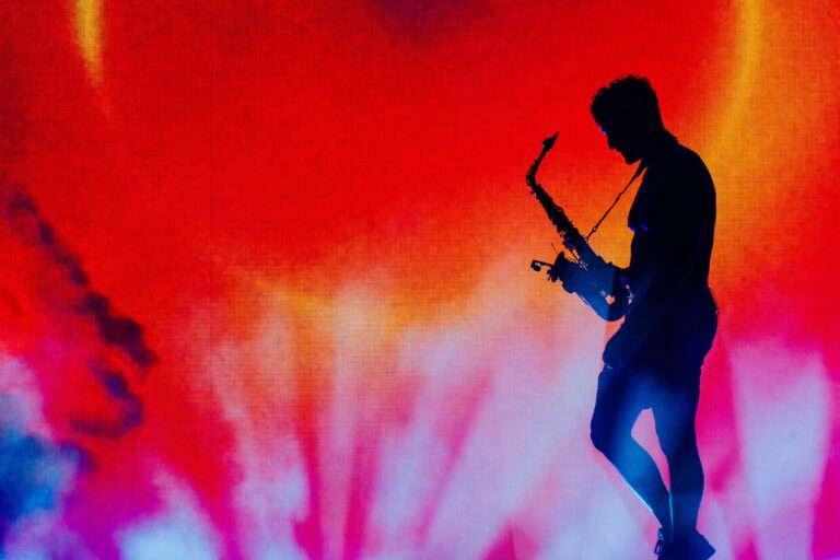 GRiZ in silhouette playing saxophone on stage against a red, orange and pink sunburst LED backdrop with blue haze at Bonnaroo 2026