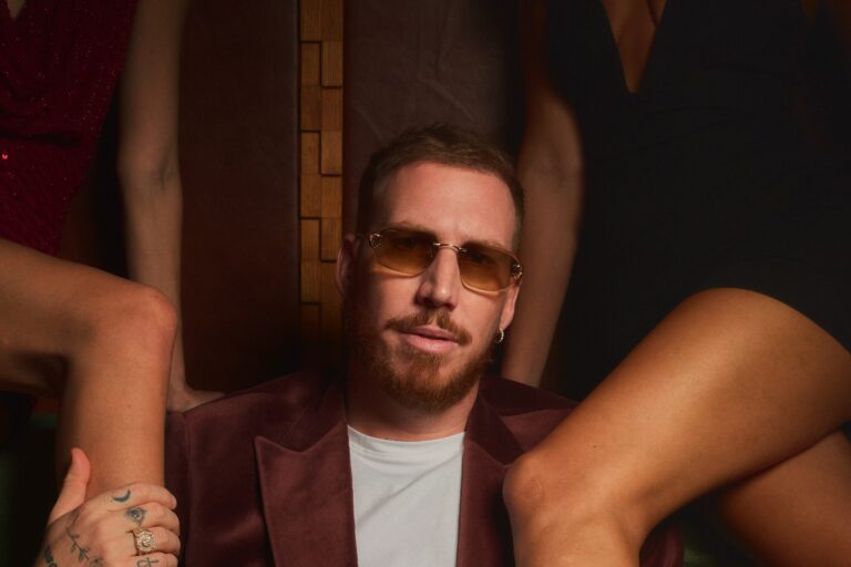 HUGEL in a maroon velvet blazer and amber-tinted sunglasses seated in a dim booth, flanked by the legs and arms of several women in cocktail dresses