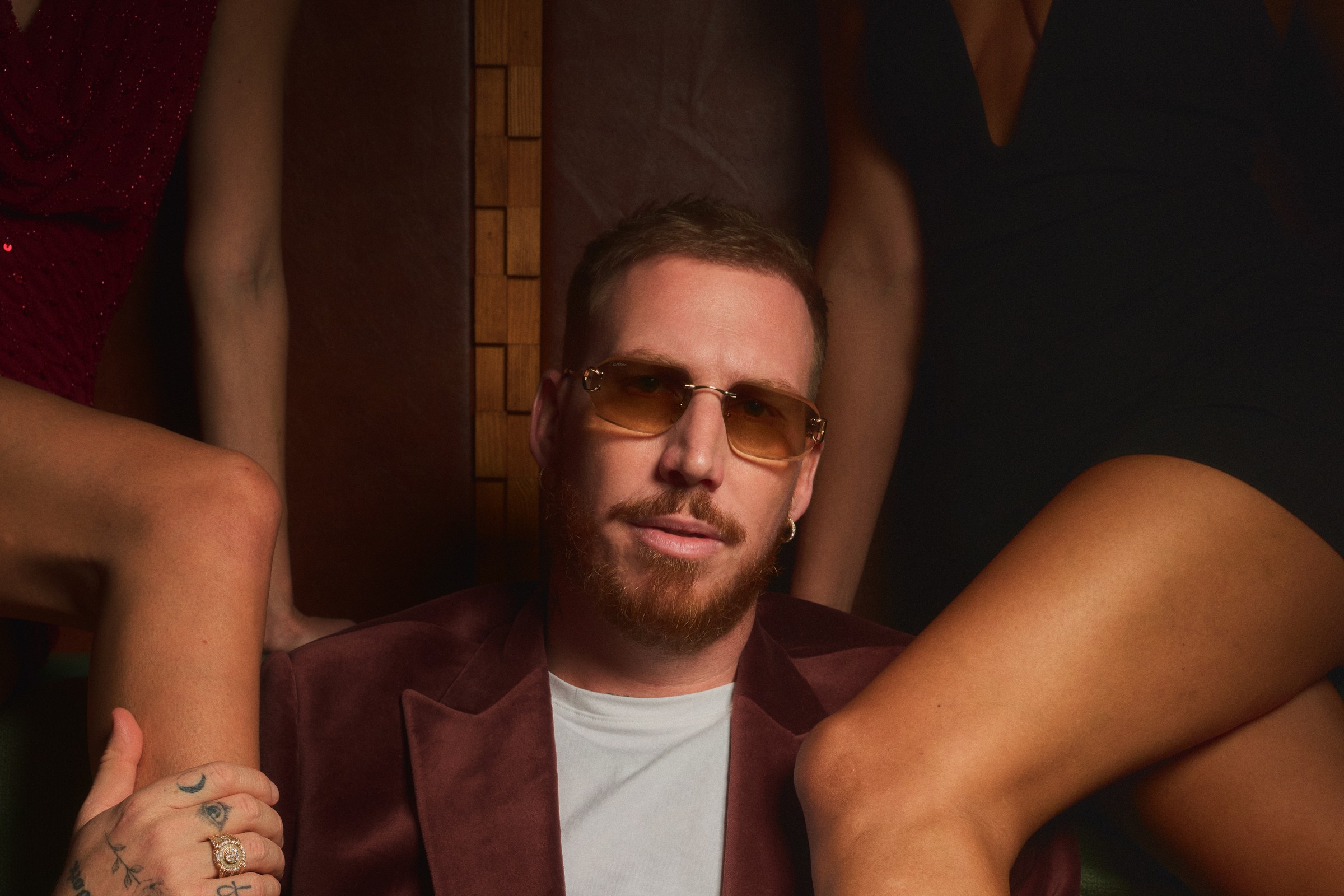 HUGEL in a maroon velvet blazer and amber-tinted sunglasses seated in a dim booth, flanked by the legs and arms of several women in cocktail dresses