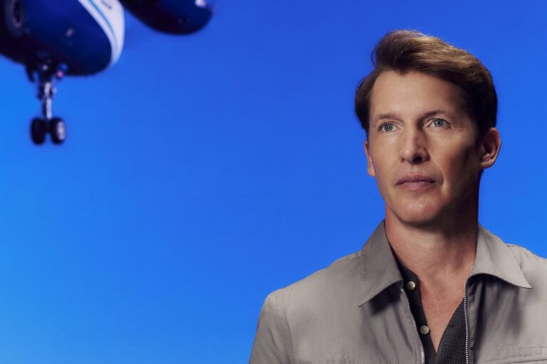James Blunt in a grey zip-front shirt over a dark polo, looking off-camera against a bright blue backdrop with a model aeroplane suspended in the upper left of the frame