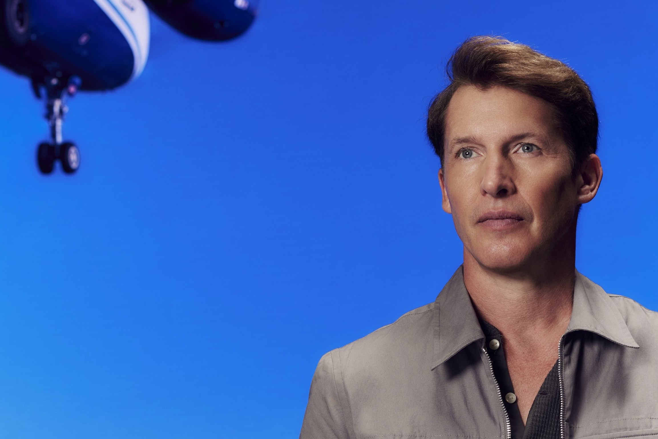 James Blunt in a grey zip-front shirt over a dark polo, looking off-camera against a bright blue backdrop with a model aeroplane suspended in the upper left of the frame