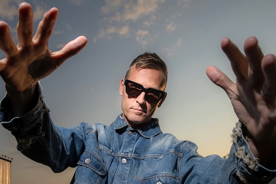 Kaskade press portrait in a distressed denim jacket and black sunglasses, both hands reaching toward the camera against a dusk sky