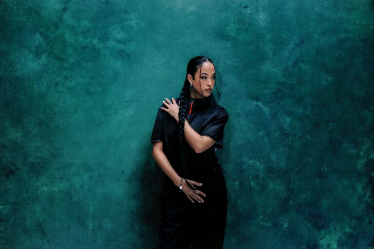 Jazzy standing against a mottled deep-teal wall in an all-black short-sleeved outfit, holding her long braid over one shoulder and looking off to the side