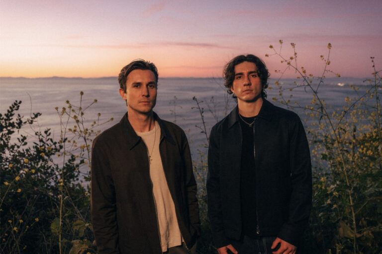 Forester's Xander Carlson and David Parris standing in coastal brush at dusk, ocean and pink sunset sky behind them, both in dark jackets