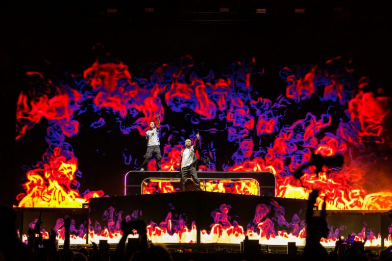 Two Friends' Matt Halper and Eli Sones standing on top of their DJ booth with arms raised, backlit by a giant LED wall of red, purple and yellow flame graphics, with silhouetted crowd hands in the foreground