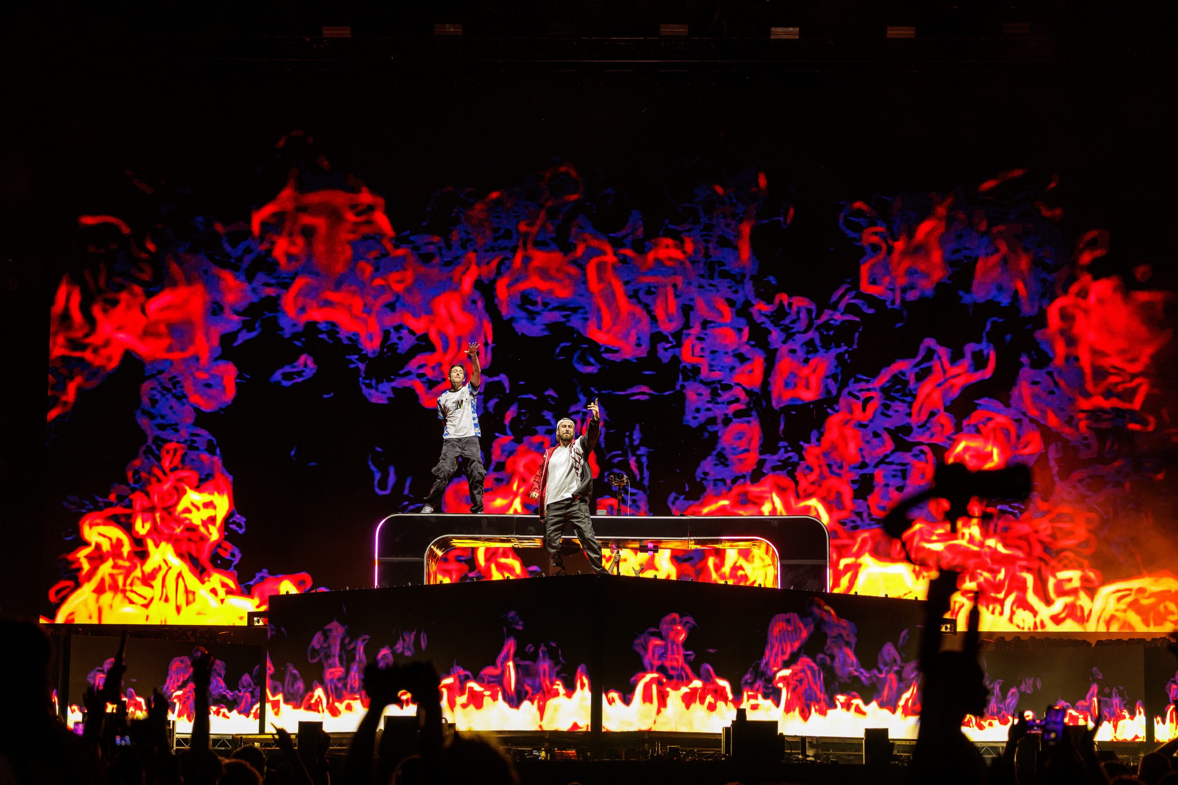 Two Friends' Matt Halper and Eli Sones standing on top of their DJ booth with arms raised, backlit by a giant LED wall of red, purple and yellow flame graphics, with silhouetted crowd hands in the foreground